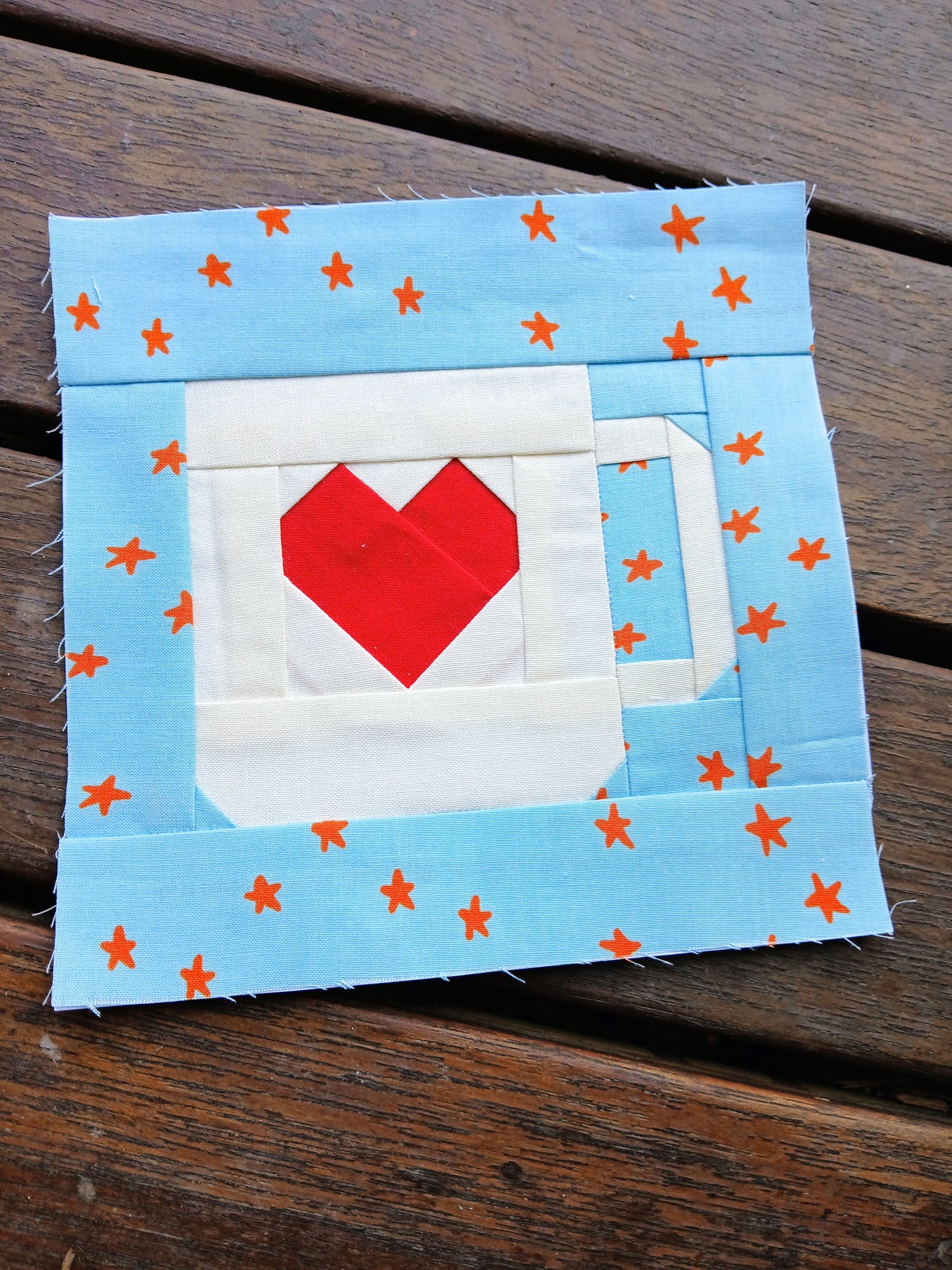 Fabric patch with a red heart and white mug on a blue background with orange stars, placed on a wooden surface.